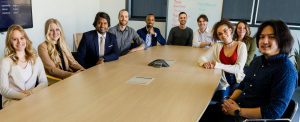 A large group of staff sitting around a meeting table, looking expectantly into the camera.