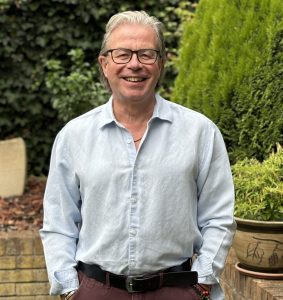 Tim Davies standing in his garden, smiling, dressed in a casual business shirt and trousers, with hands in pockets.