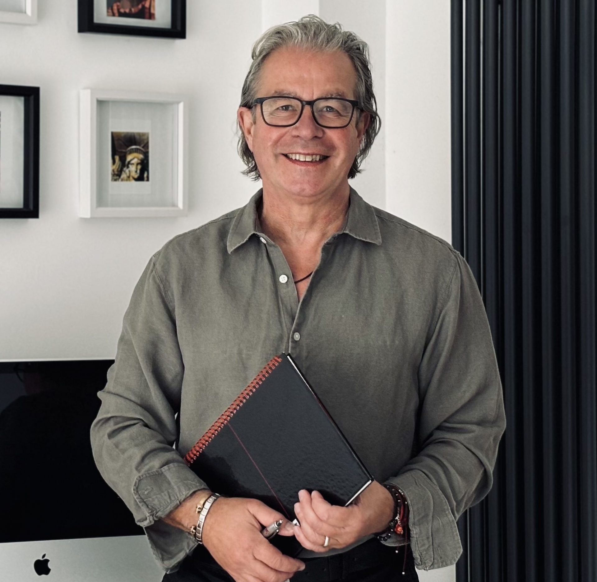 Tim Davies smiling, standing in front of some small pictures and a Mac, dressed in a casual business shirt, holding a notebook.