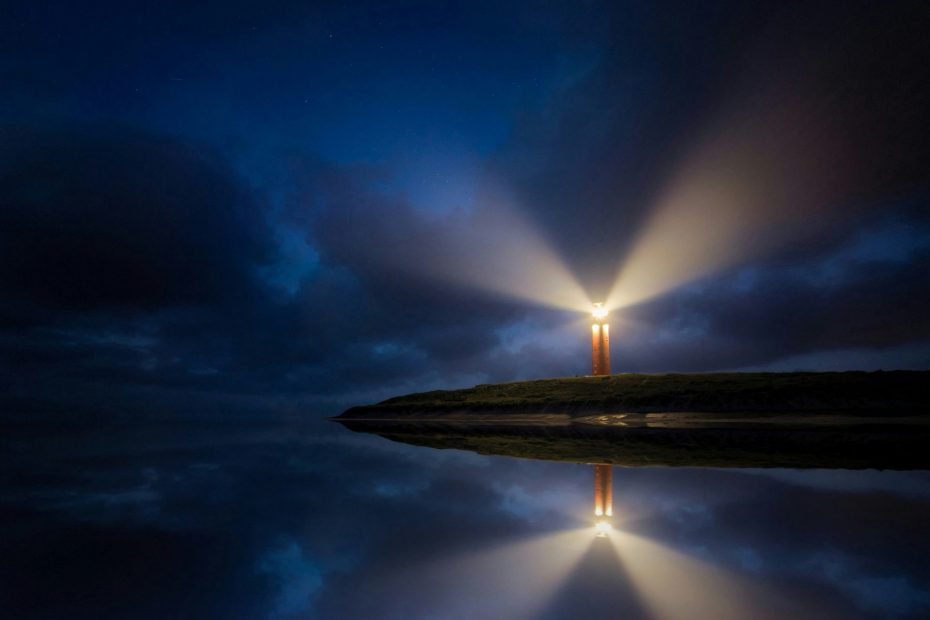 Lighthouse in distance at night with lamp shining brightly and reflected in sea.