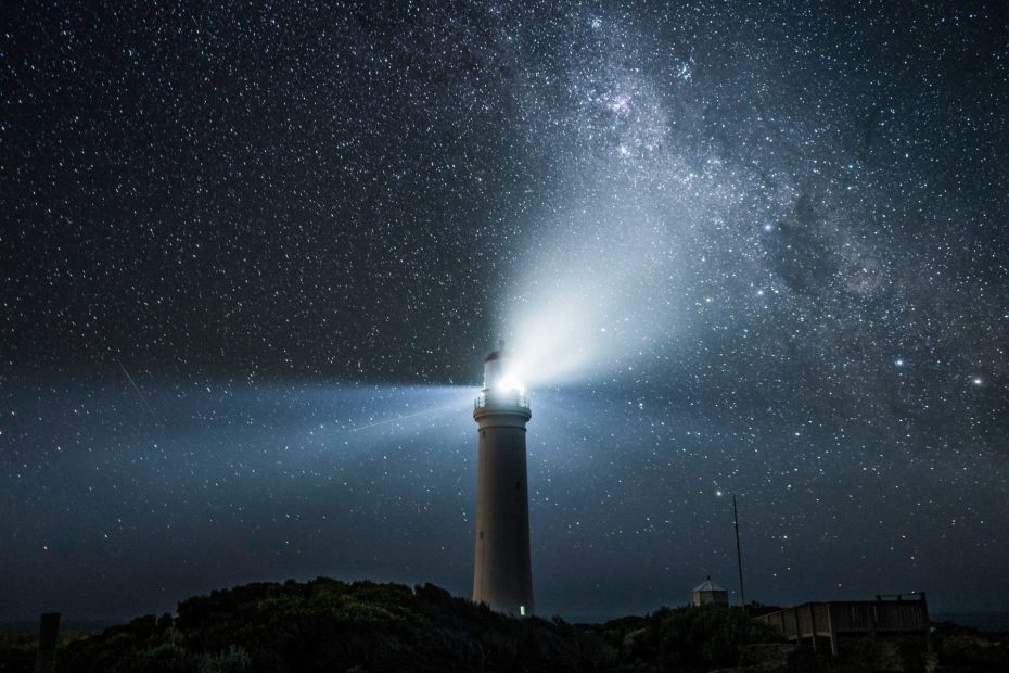 Lighthouse at night with lamp lighting up a star-filled sky.